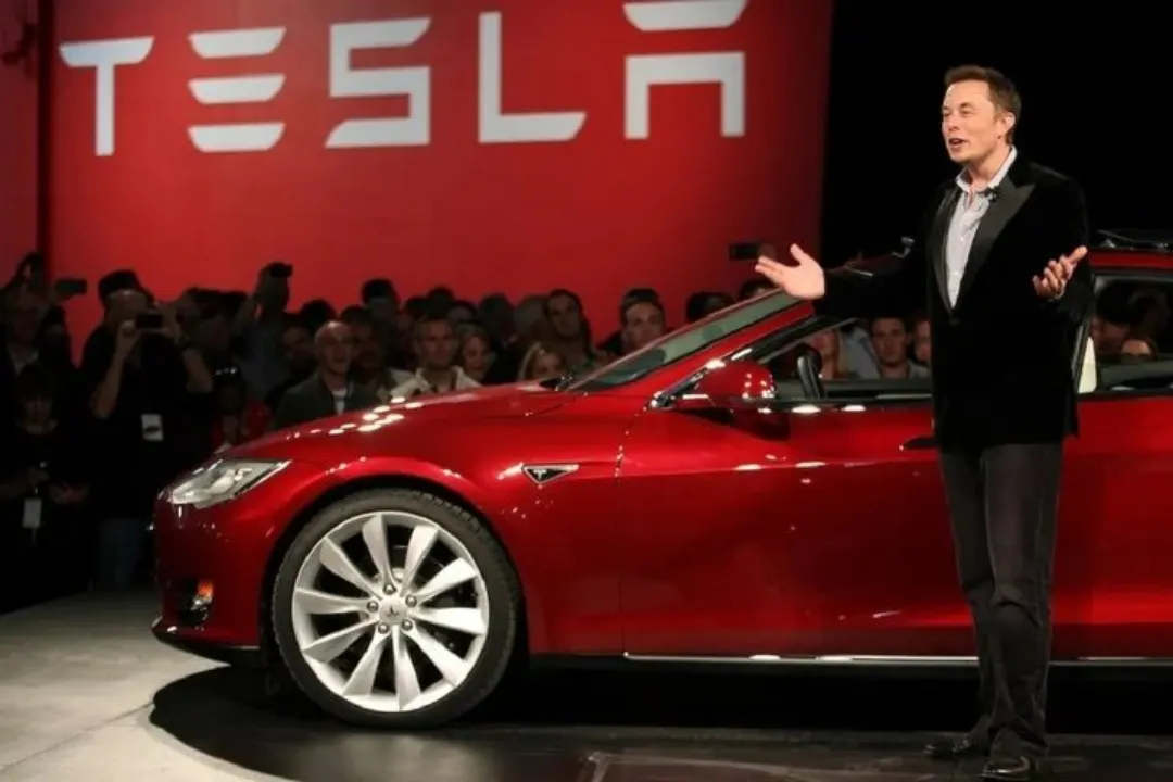 Tesla lansmanında sahnede konuşan Elon Musk, arkasında kırmızı araç ve Tesla logosu