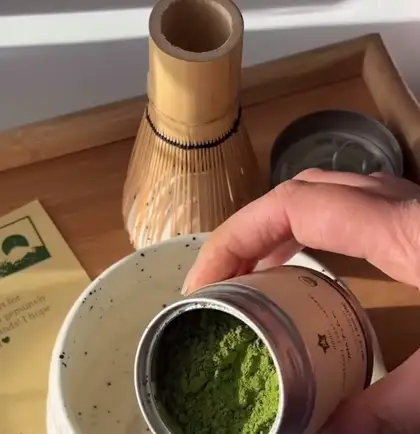 Kutu açılıyor, matcha tozu seramik kâseye dökülüyor, yanında bambu çırpıcı